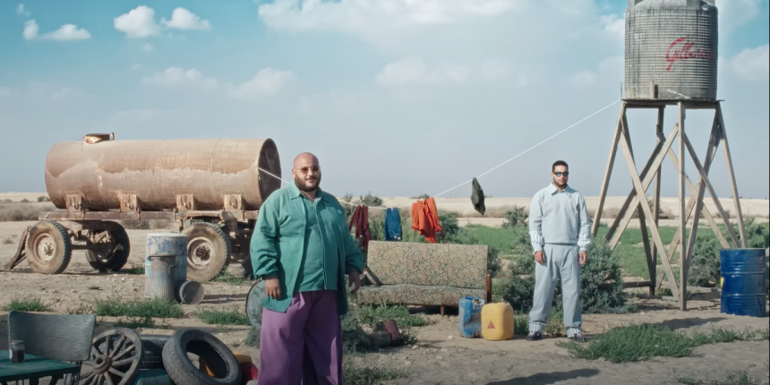 ahmed basyoni wearing purple flowy pants a turquoise jacket and shirt is standing on a muddy field w